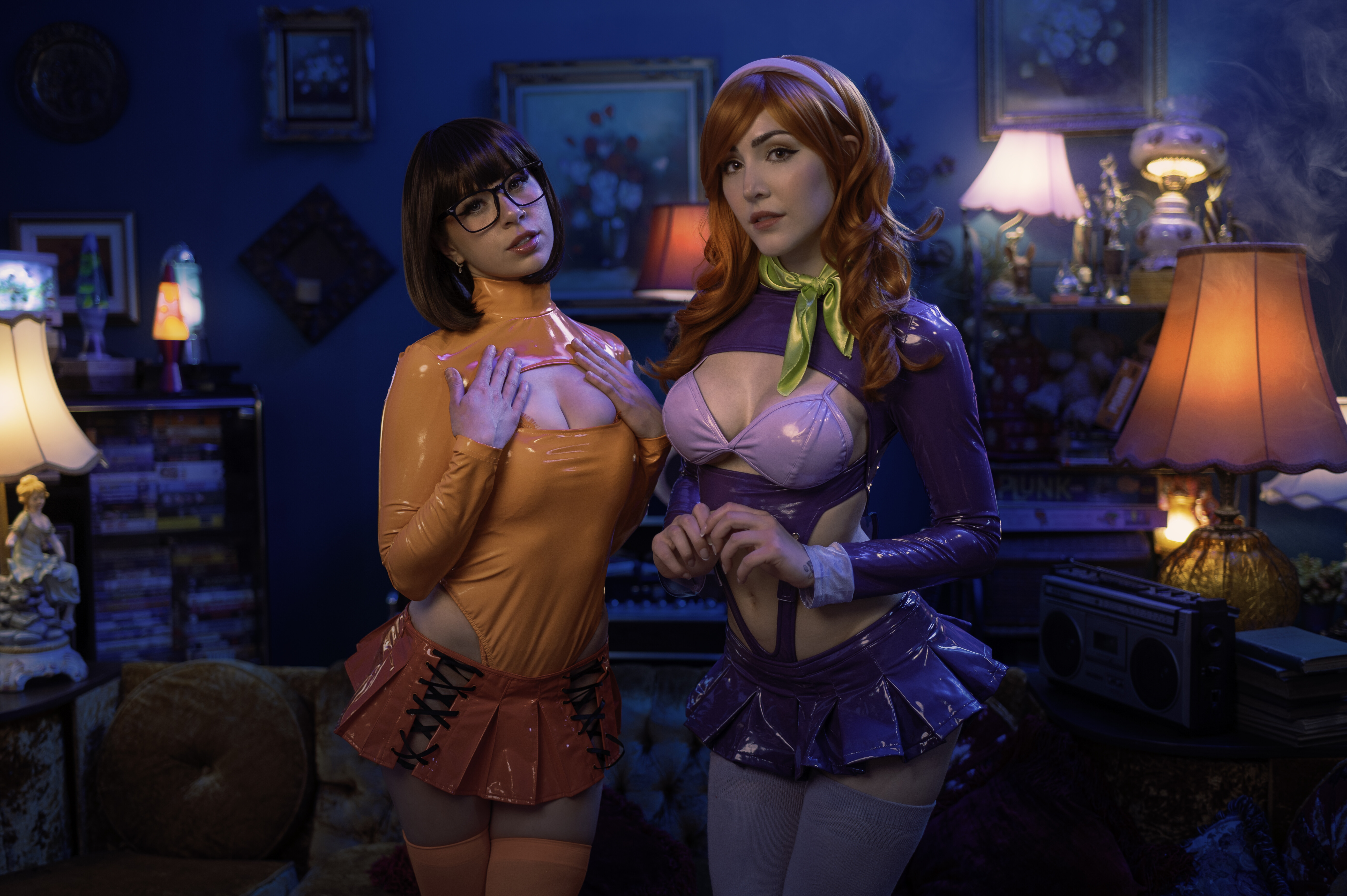 Two cosplayers in glossy orange and purple vinyl Mystery Inc. costumes stand side by side in a moody blue parlor lit by lava lamps and a boombox, one in a bob wig and black glasses, the other in a lavender headband and lime scarf.
