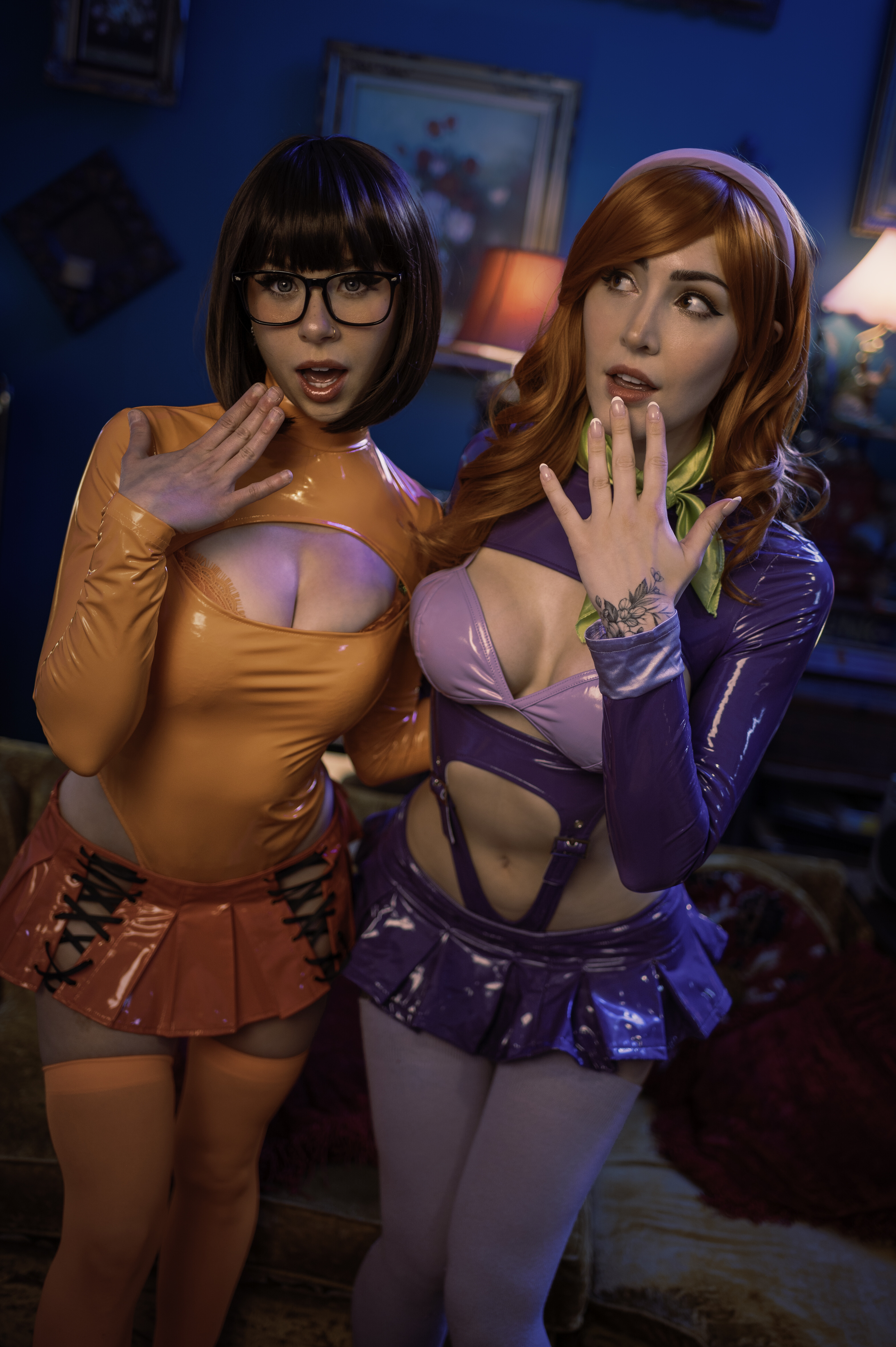 The two cosplayers lean together and raise their hands in matching gasps, lit in deep blue and violet.