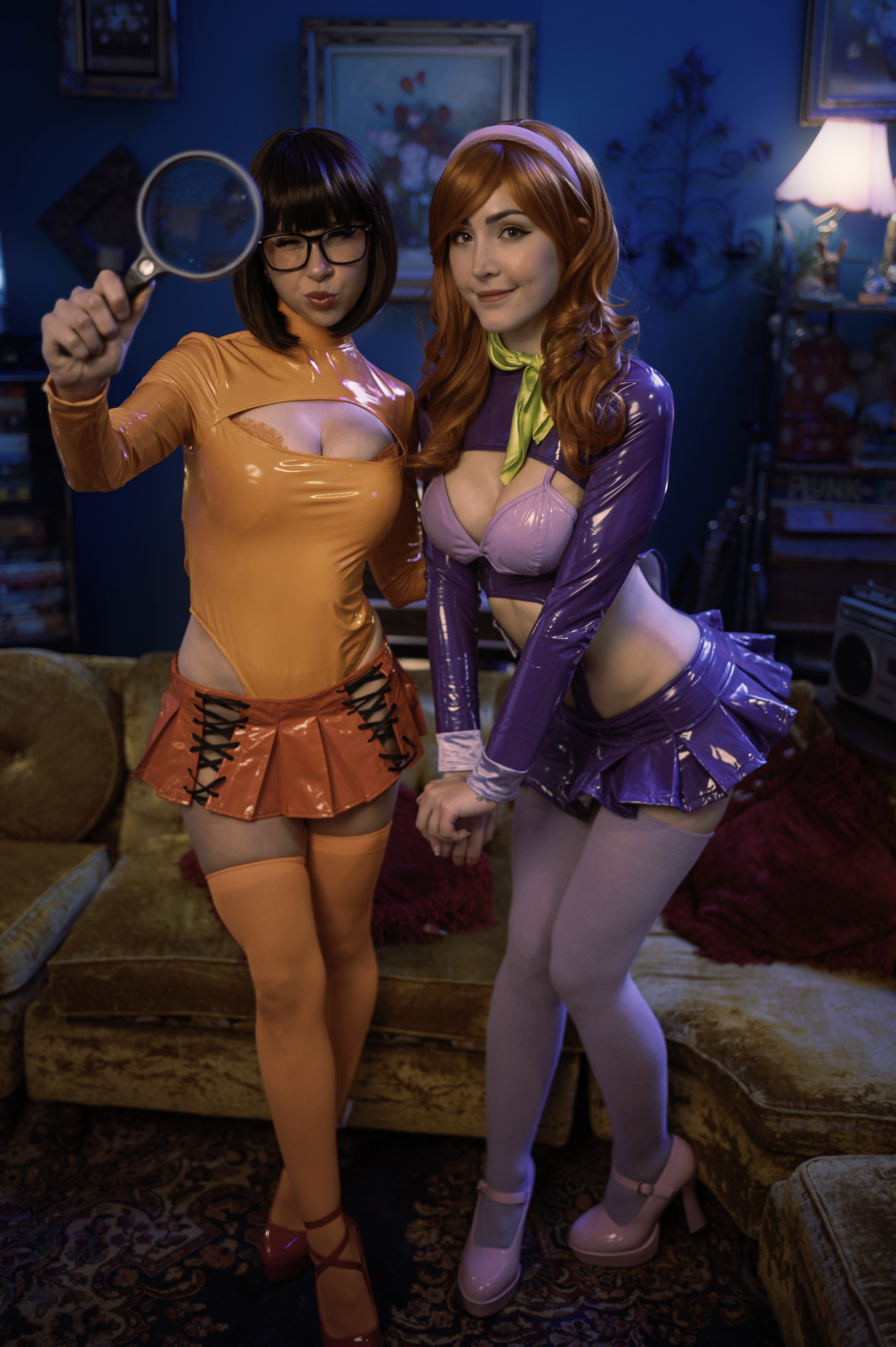 One cosplayer winks through a magnifying glass while the other in purple vinyl and lavender stockings leans in with a knowing smile.