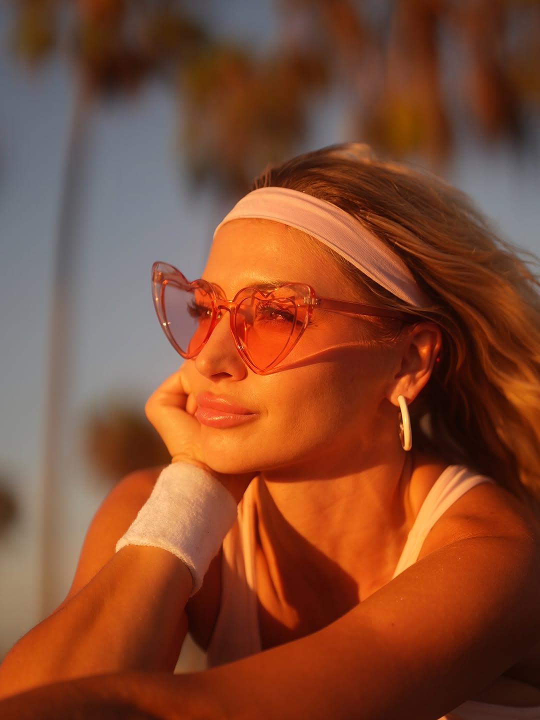 Close-up portrait of Katerina in heart-shaped pink sunglasses and a white headband, resting her chin on a wristband