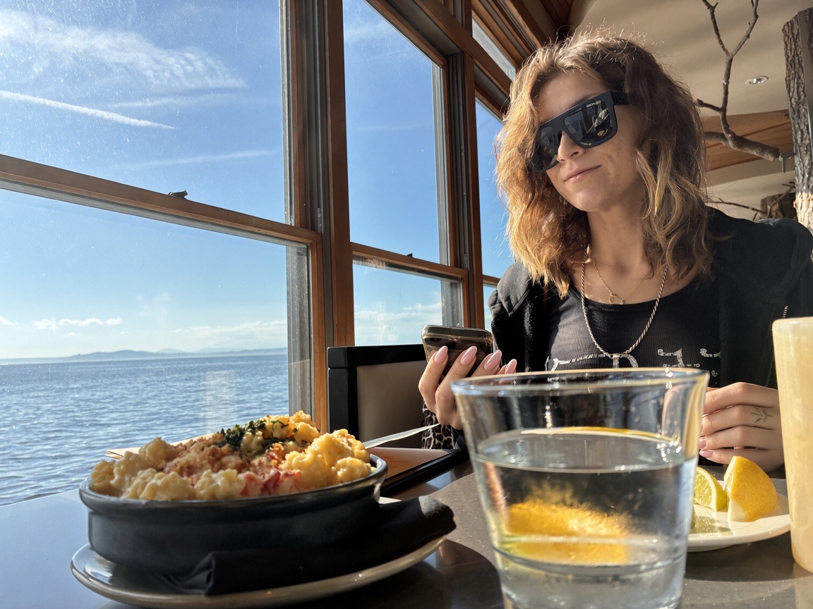 Siarra Cat at the window seat looking at her phone beside lobster mac and a water glass with lemon, Elliott Bay behind her