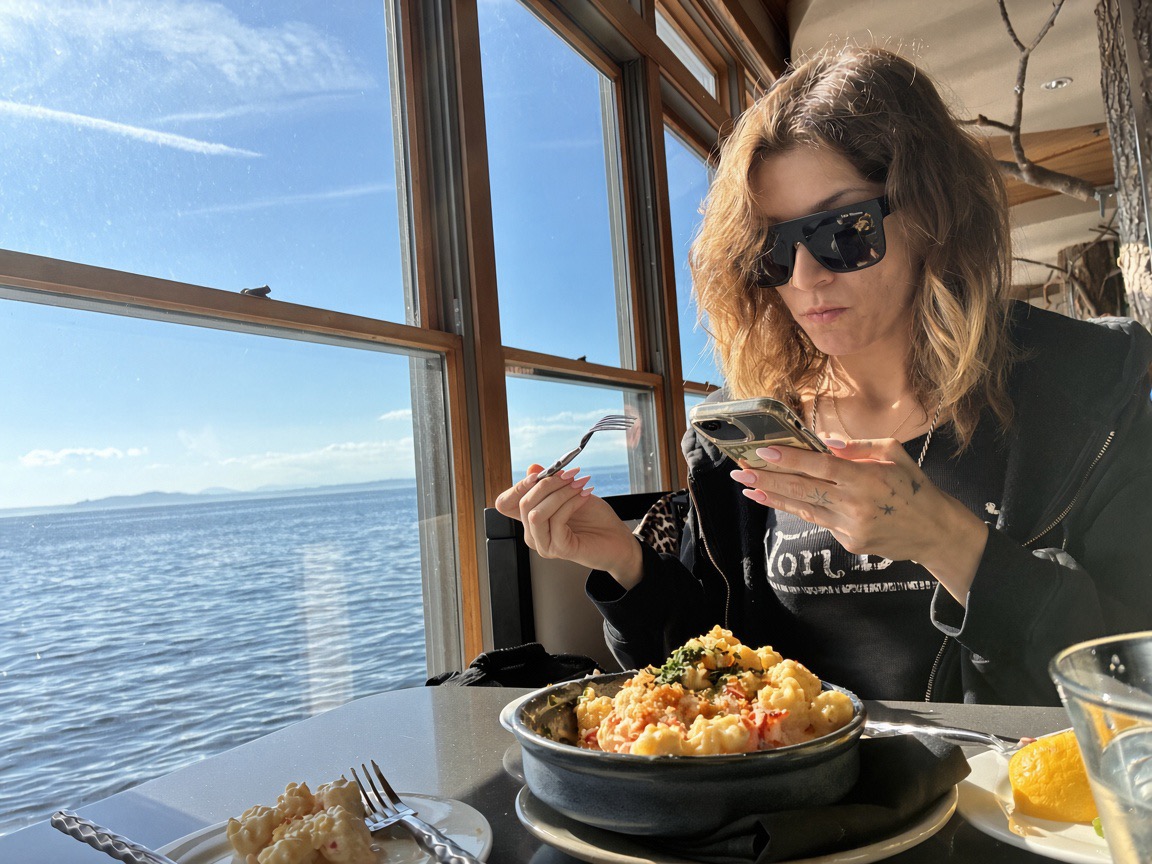 Siarra Cat checking her phone with a fork in the other hand, lobster mac in front of her, at the window view