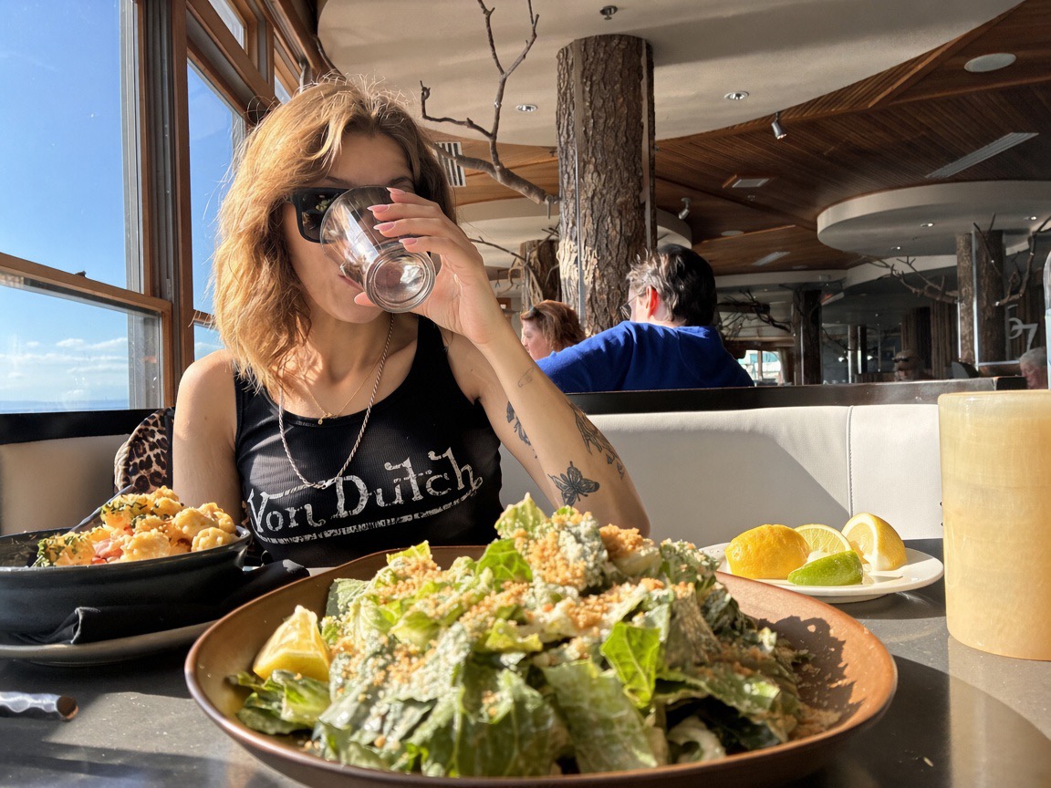 Siarra Cat drinking from a water glass at Six Seven with Caesar salad in the foreground and lobster mac and citrus on the table